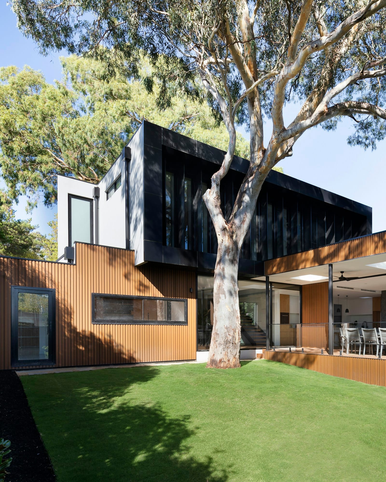 A low villa in dark cladding and timber, set behind a lawn and a mature tree